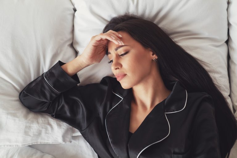 Tanned woman with black hair lying on a bed in black pajamas, appearing to be sad and lost in thought.