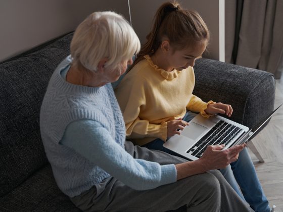 An elderly woman holding a computer open for a young girl in a yellow shirt.